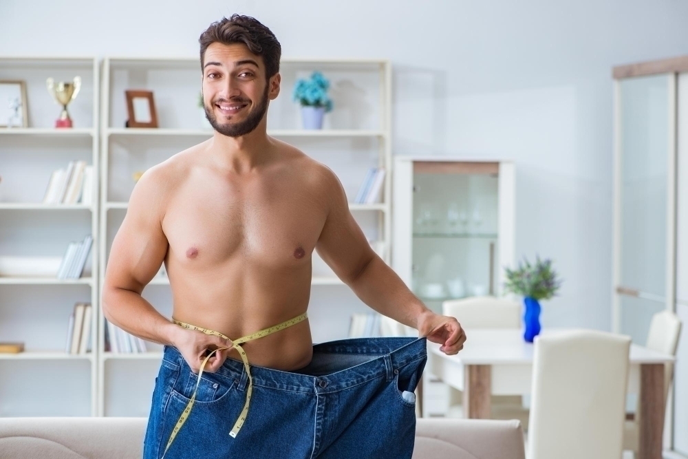 A joyful man showcasing his weight loss by pulling his loose jeans at the waist in a bright modern home