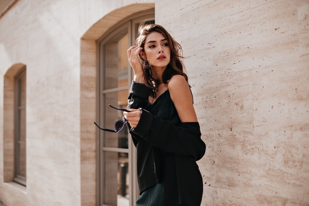 Cute young girl with dark wavy hairstyle and bright makeup silk dress black jacket holding sunglasses in hands and looking away against beige building wall2